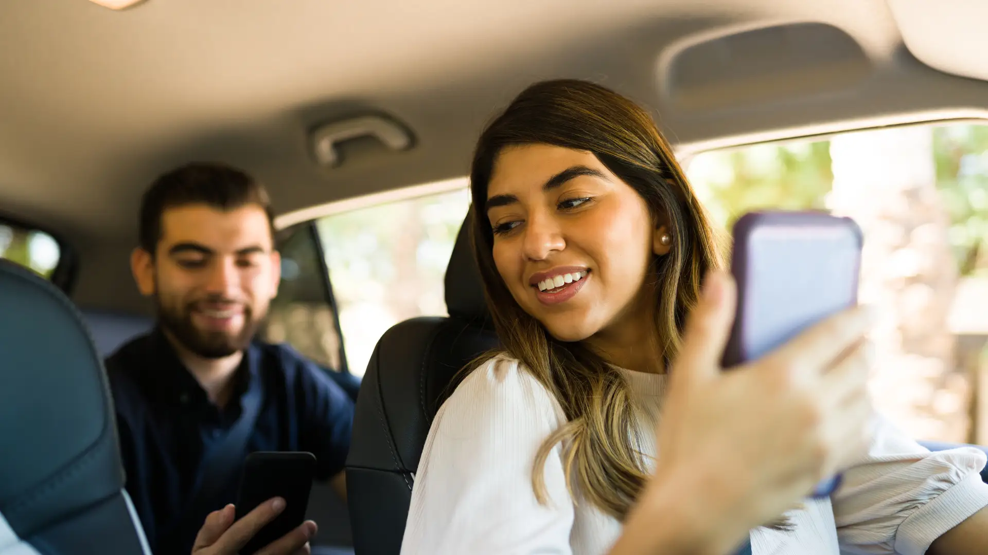 A woman sitting in a car, smiling and taking a selfie with a phone, while a man in the background is also using his phone