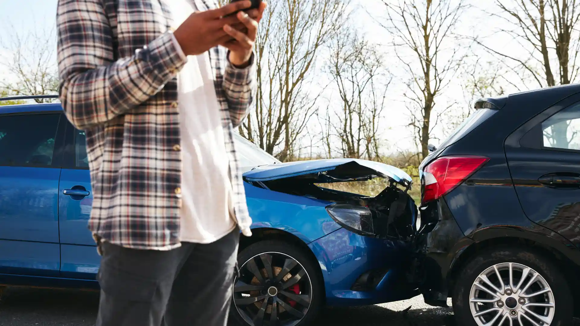 Person using a phone near the scene of a rear-end car accident involving two vehicles, one with visible front-end damage.