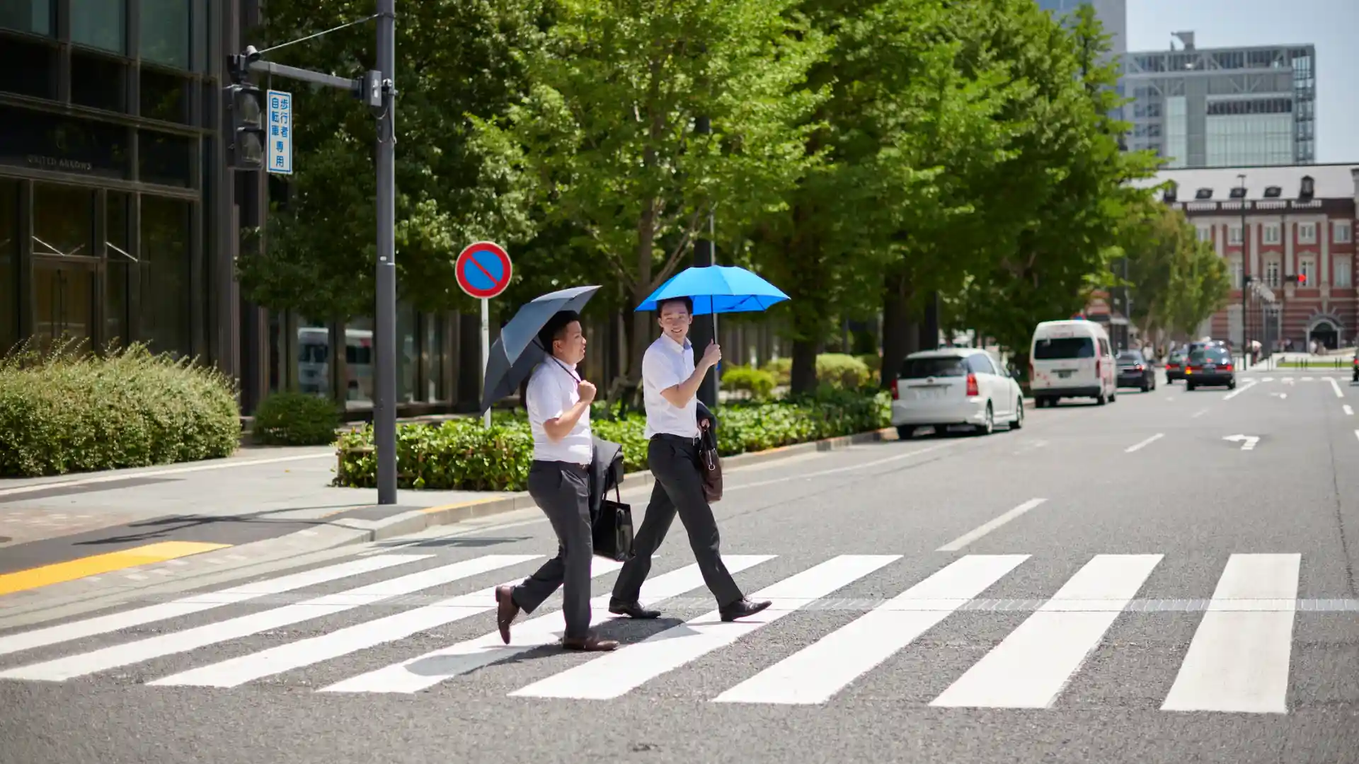 Two people crossing a street at a crosswalk while holding umbrellas on a sunny day.