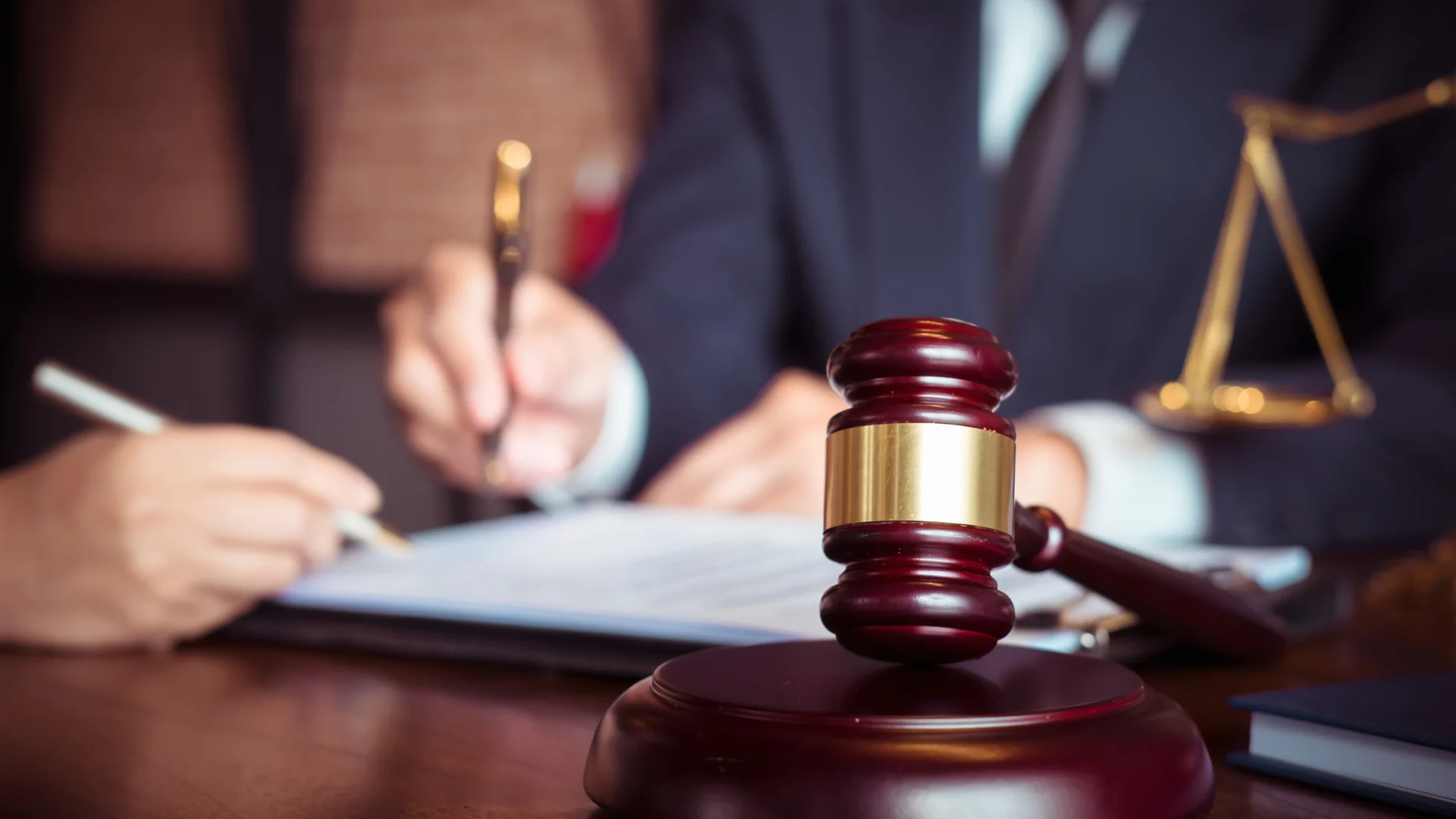 A close-up of a wooden gavel on a desk with a person signing legal documents in the background, representing legal proceedings