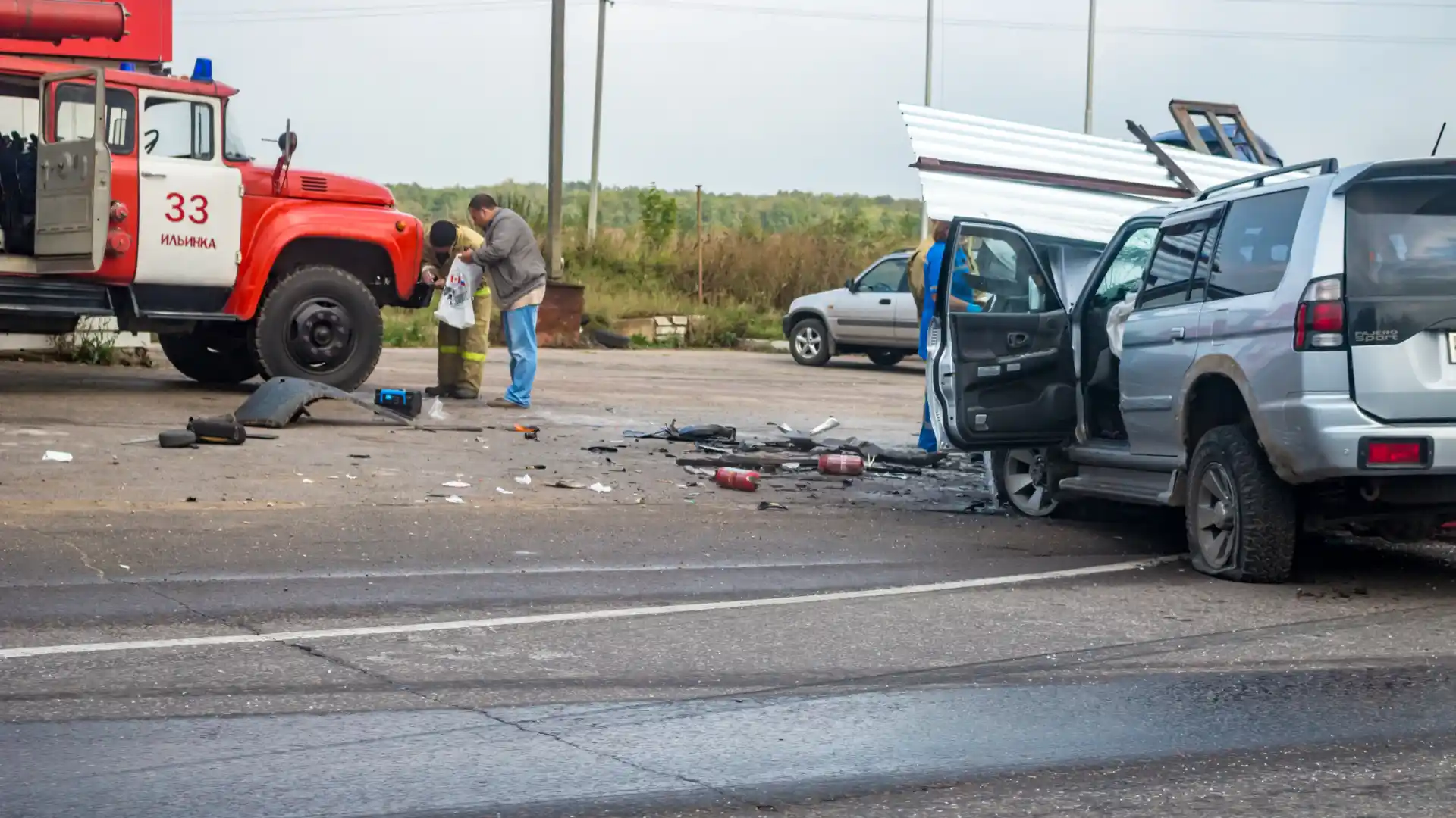 A car accident scene with a person inspecting the damage while another vehicle is seen nearby, with scattered debris on the road.
